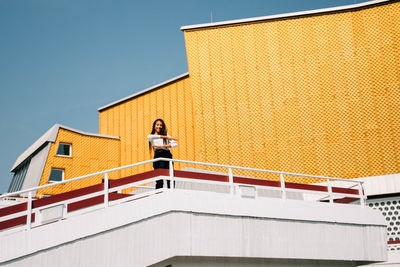 Low angle view of young woman standing by building against sky