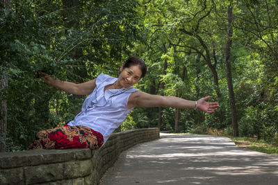 Cheerful senior woman with arms outstretched sitting on retaining wall in forest