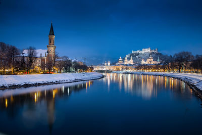 Illuminated buildings by river against blue sky