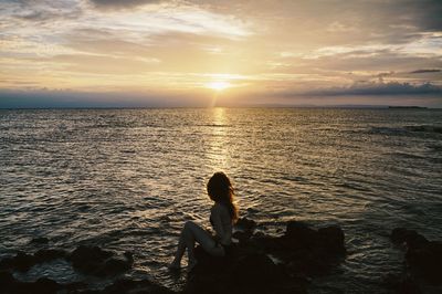 Woman sitting on rock by sea against sky during sunset