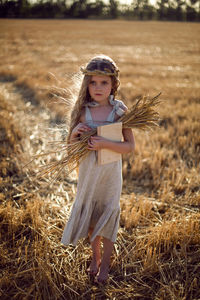 Girl child in a dress and a wreath on her head stands on a mown field of wheat at sunset in summer