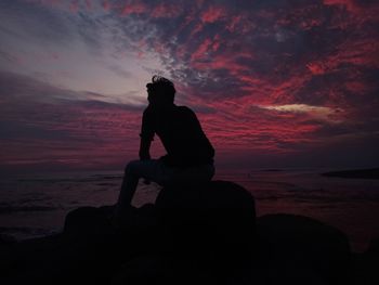 Silhouette man looking at sea while sitting on rock against sky during sunset