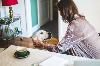 Woman stroking dog while having coffee on dining table at home