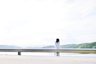 Rear view of woman sitting on railing by lake against sky