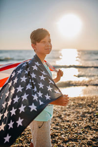 Man holding flag while standing at beach against sky during sunset