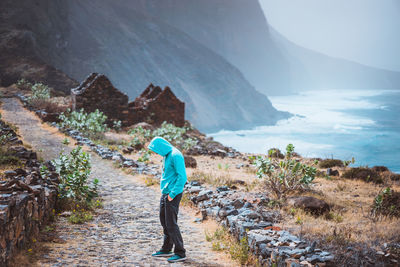 Side view of man wearing blue hooded short standing by mountain and sea