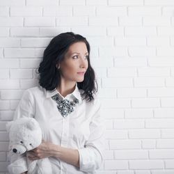 Thoughtful woman holding teddy bear while standing by white brick wall