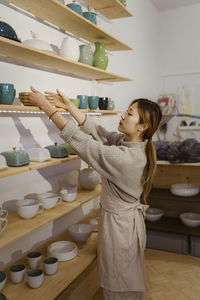 Side view of young female potter arranging merchandise on shelf at workshop