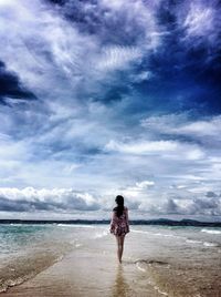Rear view of woman standing on shore at beach against cloudy sky
