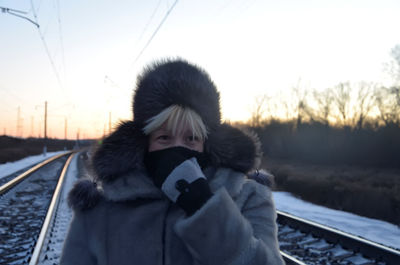 Woman wearing warm clothing and fur hat standing on railroad against sky during sunset