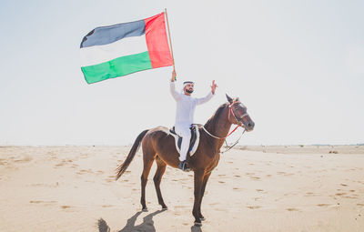 Man holding flag and riding horse against sky