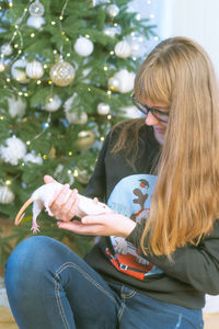 Girl holding rat while sitting against christmas tree