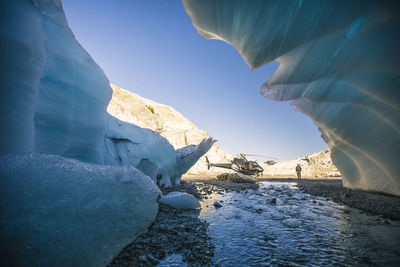 River flows out of glacier toward parked helicopter