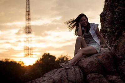 Young woman sitting on rock against sky during sunset