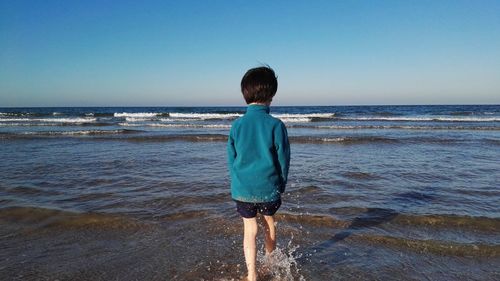 Rear view of boy walking at beach against clear blue sky