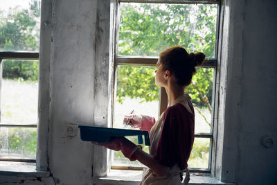 Side view of woman using phone while standing on window