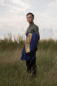 Portrait of young woman holding graduation gown while standing on field against sky