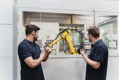 Male coworkers looking at robotic arm through window while standing in factory