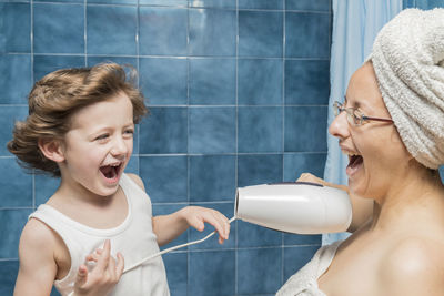 Cheerful mother blowing air with dryer on son face in bathroom