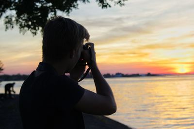 Side view of man photographing sea against sky during sunset