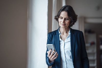 Businesswoman holding smart phone while leaning on wall at home office