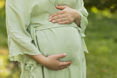 Cropped pregnant woman puts,holds hands on big belly, green trees,meadow on background