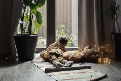 Boy looking out of window lying on rug with stuffed toy at home