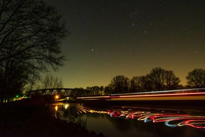 Light trails by river against sky at night