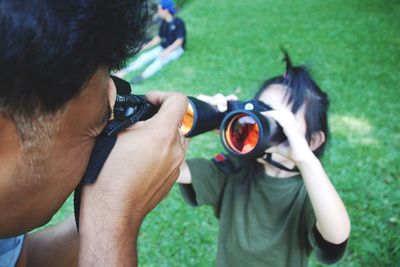 Father photographing girl looking through binoculars while standing on field