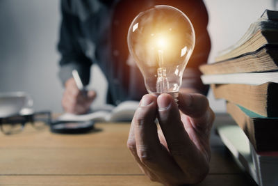 Midsection of man holding illuminated light bulb by stacked books on table