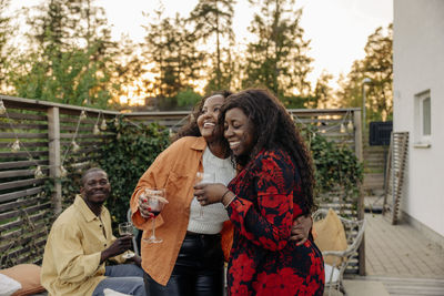 Smiling woman embracing female friend while holding drinks at dinner party