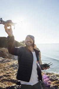 Smiling man flying quadcopter while standing at beach against clear sky during sunny day