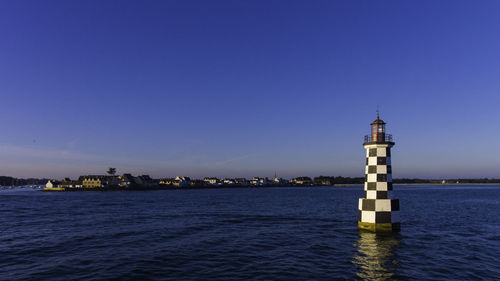 Lighthouse by sea against clear blue sky