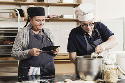 Woman holding digital tablet while coworker cooking food in kitchen