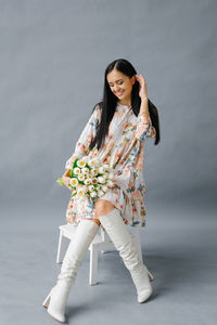 A young pretty woman in a dress and white boots sits on a white chair with a bouquet of white tulips