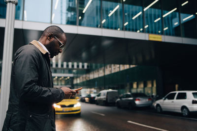 Side view of businessman using smart phone while standing on road against building in city