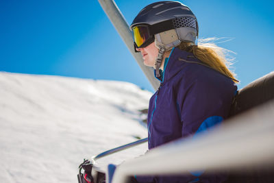 Woman wearing ski goggles and helmet while sitting in ski lift against sky