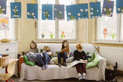 Full length of boy and girls reading books while sitting on sofa in classroom