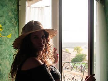 Portrait of woman wearing hat while standing against window at home