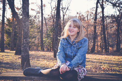 Happy girl sitting on bench against bare trees in forest
