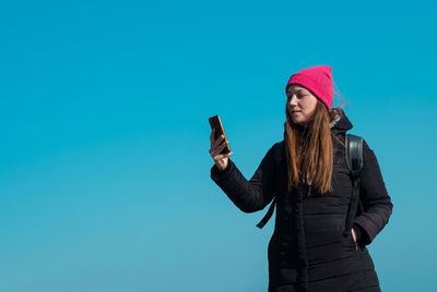 Young confident woman in pink hat smartphone in hand calling friend walking on blue sky background.