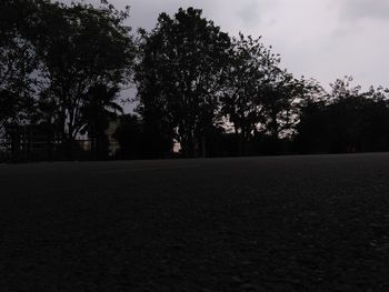 Empty road amidst silhouette trees on field against sky