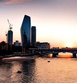 Modern buildings by river against sky during sunset