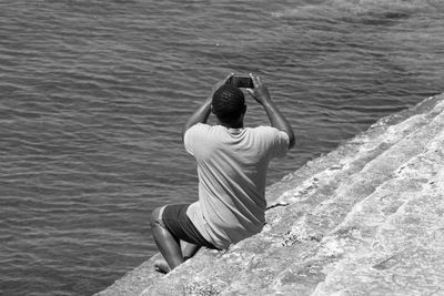 Rear view of man photographing through mobile phone while sitting on steps by sea