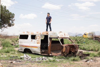 Man standing on abandoned car on field against sky