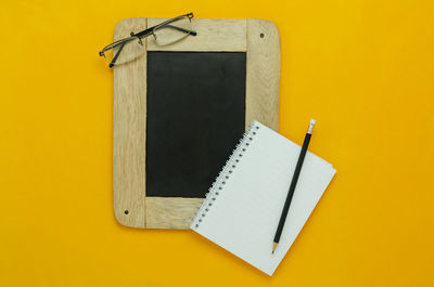 Directly above shot of blackboard with spiral notebook and pencil on yellow background