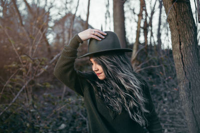 Smiling woman with long hair holding hat while standing against trees in forest