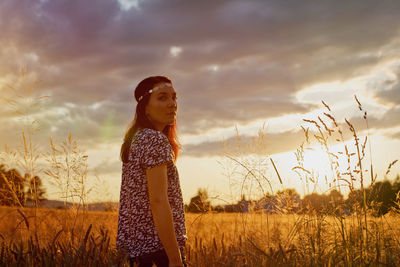 Young woman standing on wheat field against cloudy sky during sunset