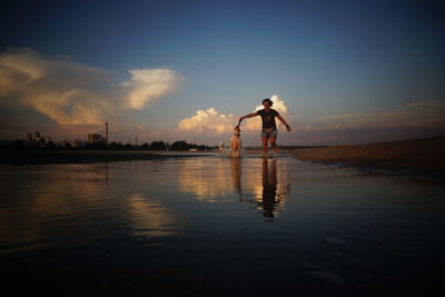 Woman with dogs running on shore at beach against sky during sunset