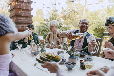 Happy friends of lgbtq community drinking wine while sitting at table during dinner party in back yard